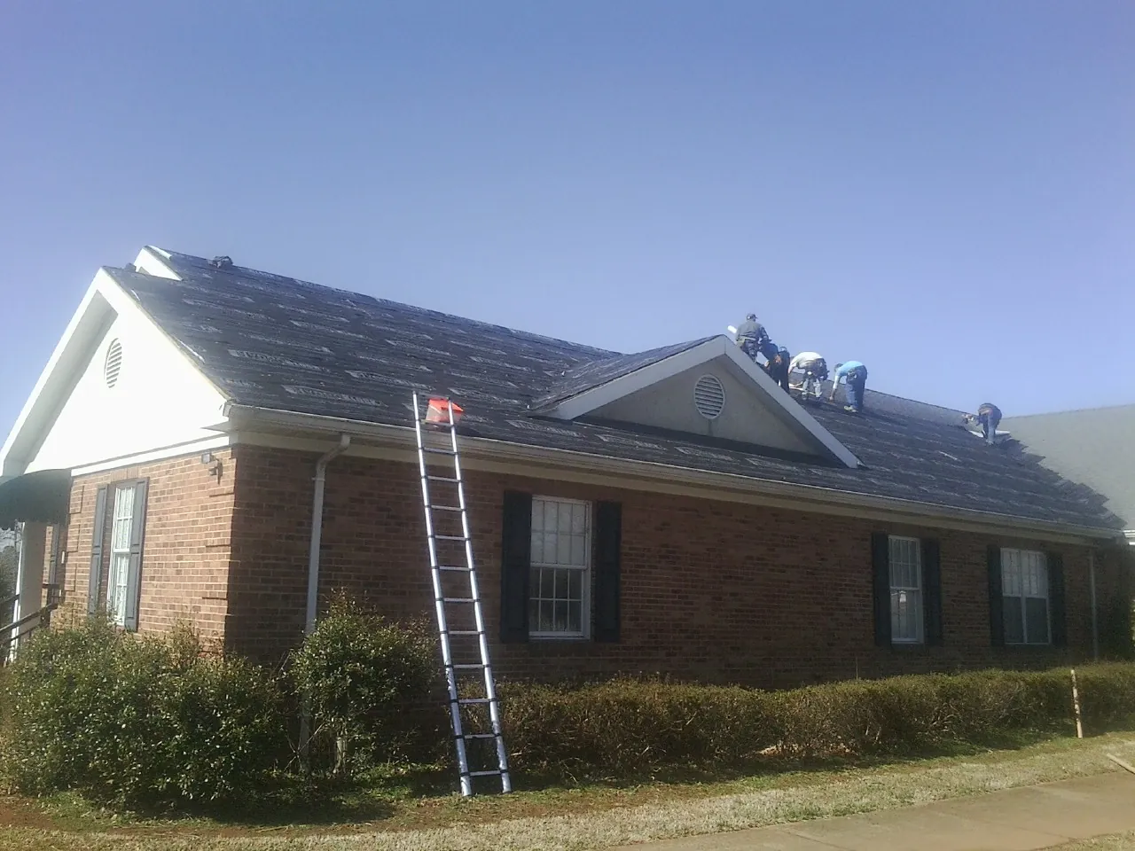 Crew tearing shingles off a brick ranch roof with a ladder set — Metal Roof Repair work by our crew in Fairport