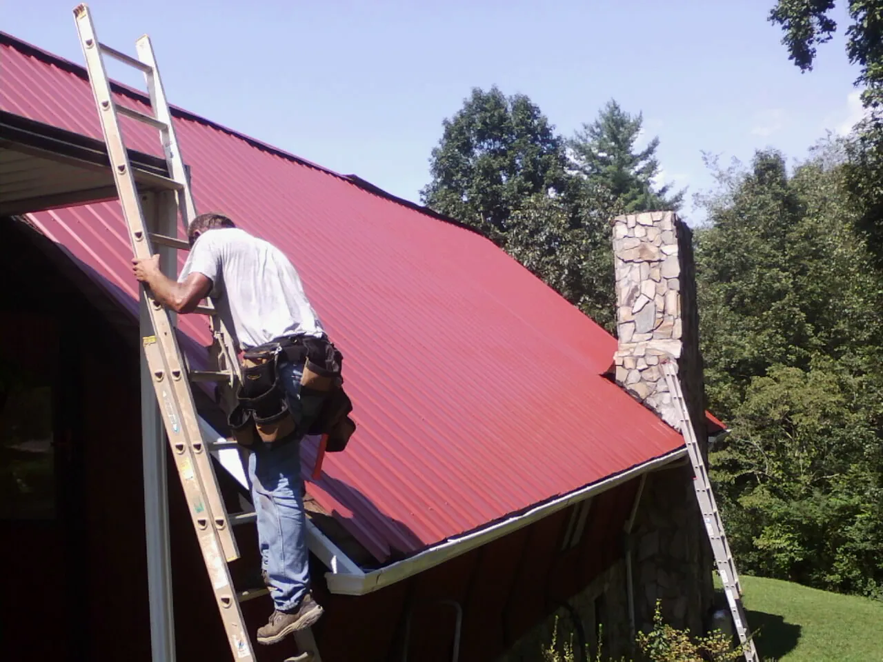 Roofer on a ladder beside a red metal roof and stone chimney — Soffit & Fascia Repair by Roofing Brothers in Fairport